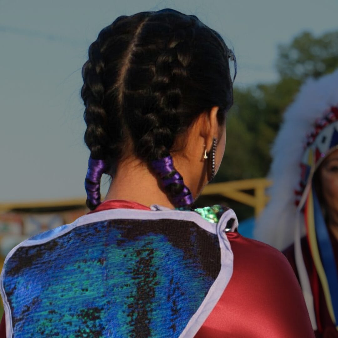 A young Indigenous girl with braided hair tied with purple ribbons, facing an older woman in the background, wearing traditional regalia.